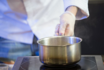 Cook's hands in white gloves hold a saucepan over an electric st