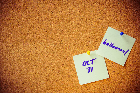 blue handwriting on white paper note, pin on cork board with yellow and blue pin, "halloween reminder" concept