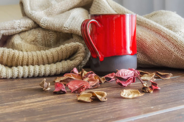 Red mug of herbal tea, beige cozy knitted sweater and dry leaves on  the wooden background. Delicious cold weather beverage for fall times. Stay at home concept.