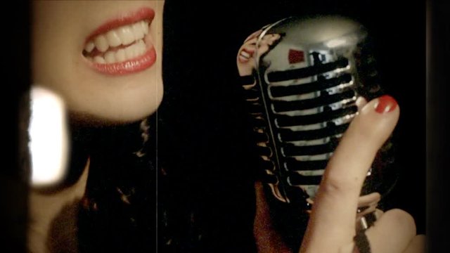 Fake 8mm amateur film: a classy young woman singing on stage with a retro vintage microphone. Making cool dance moves. Extreme close-up shot, detail of mouth, lips and lipstick, black background.
