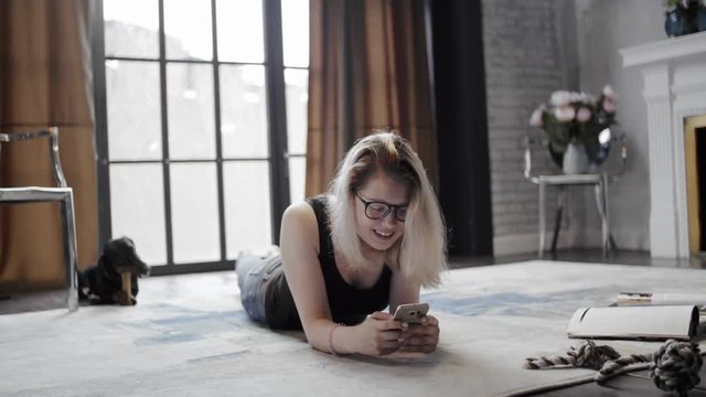 Girl in glasses with mobile phone lying on floor on background dachshund dog