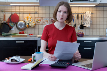 Thoughtful stressed young female sitting at kitchen table with papers and laptop computer trying to work through pile of bills, frustrated by amount of domestic expenses while doing family budget