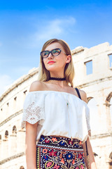 Nicely dressed woman in colorful shirt and white blouse with lot of accessories as choker, glasses, watch and bracelet. She has gorgeous long curly blonde hair.