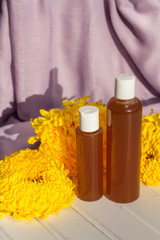 Two bottles with gel stand on white wooden table in front of yellow chrysanthemums and purple fabric.