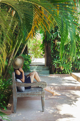 Young woman relaxing under the palm trees, hot sunny day,Bali.A young girl in a straw hat resting in the shade of coconut palms on a carved beautiful bench.Luxury holidays.