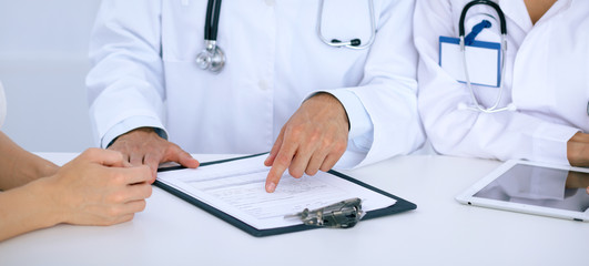 Fototapeta premium Doctor and patient talking while sitting at the desk in office. Physician pointing into medical history form. Medicine and health care concept