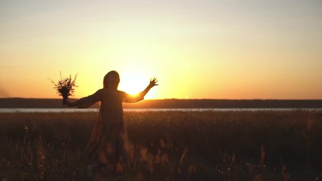 picture of lovely girl with long hair running skipping through meadow covered with plants holding bunch of flowers in hand with amazing sunset background slowmotion