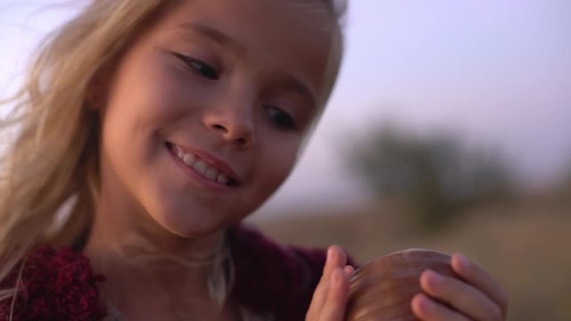 closeup smiling cheerful little girl with blond hair being excited and happy finding big beautiful shell holding it to ear listening noise in it with closed eyes in morning slowmotion 