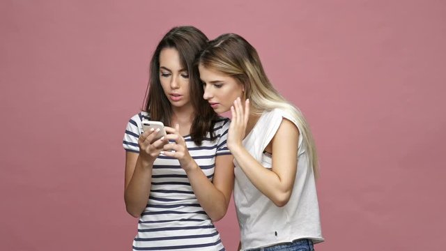 Cheerful friendly caucasian women typing on smartphone and talking isolated over pink