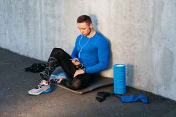 Young sports man in blue T-shirt siting on mat and listening music on the background of gray wall