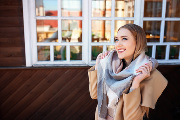 ttractive blonde woman in urban background with sun backlight. Young girl wearing winter coat and scarf standing in the street. Pretty female with straight hair hairstyle and blue eyes.