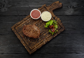 Grilled beef steak with sauces on a board. Dark wooden table.