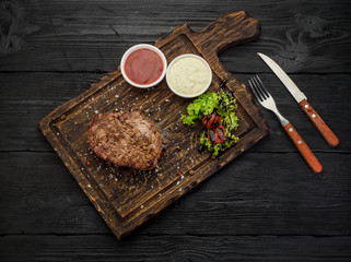 Grilled beef steak with sauces on a board. Dark wooden table.