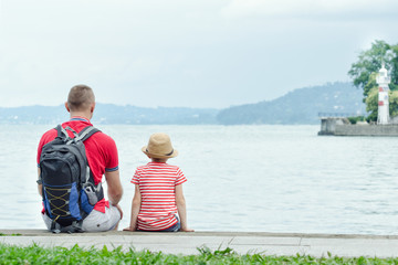 Fototapeta premium Father and son sitting on the pier on the sea background, lighthouse and mountains in the distance. Back view