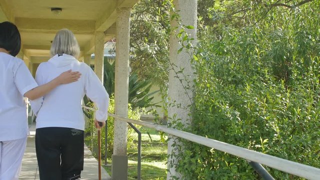 Elder woman with stick helped by nurse in rehab clinic garden.