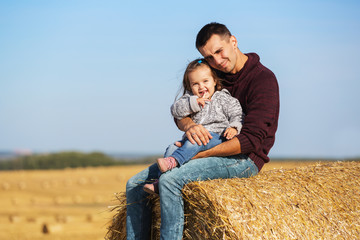 Happy father and 2 year old girl sitting on hay bale in harvested field