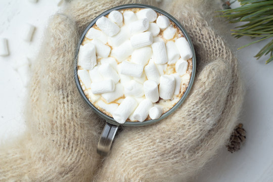 hands in white mittens holding mug with hot chocolate close-up, coffee and marshmallows, Christmas tree branches, fir cones, Christmas tree decorations and white background, c