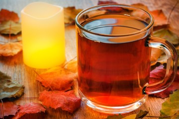 Glass cup of tea and glowing candle on wooden table with autumn leaves.