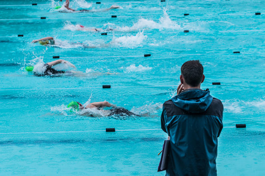 Male Swimming Coach Standing By The Swimming Pool In The Rain Watching Swimmers Racing By