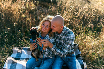 man guy in checkered shirt and girl in green dressed in a black shirt and jeans kiss standing in grass sitting in grass half on top of sunset with dog running around fooling around - Powered by Adobe