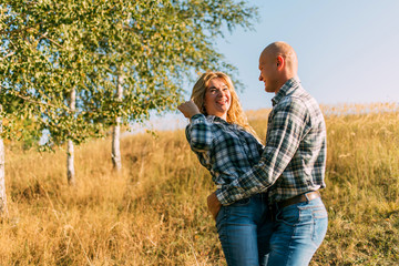 male guy in checkered shirt and girl in green dressed in a black shirt and jeans kiss standing in grass sitting in grass half on top of sunset with dog running around fooling around