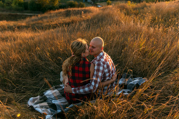 man guy in plaid shirt and woman girl in red checkered dress kiss standing in grass sitting in grass half on top on sunset background