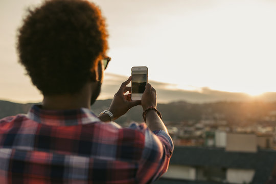 Young african american man taking a photo on his smartphone from a rooftop at sunset.