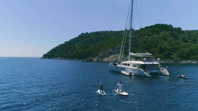 Aerial Following Shot of a Two Girls Standup Paddleboarding Near Their Sailing Yacht. Luxury White Yacht is Anchored Near Beautiful Green Rocky Coast. Shot on Phantom 4K UHD Camera.