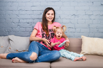 beautiful young mother uses a smartphone to play with a two-year-old daughter sitting on the couch in the room Dressed in bright colors, colored clothes