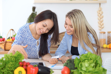 Two young happy women making online shopping for making menu by tablet computer. Friends cooking in the kitchen