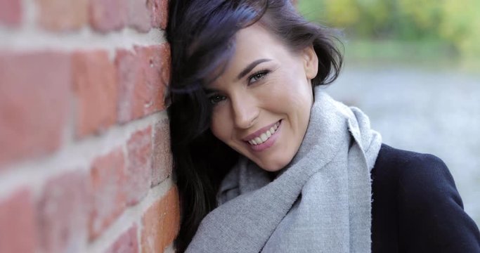 Attractive woman in black jacket and soft gray scarf leaning her head on brick wall on sunny autumn day.