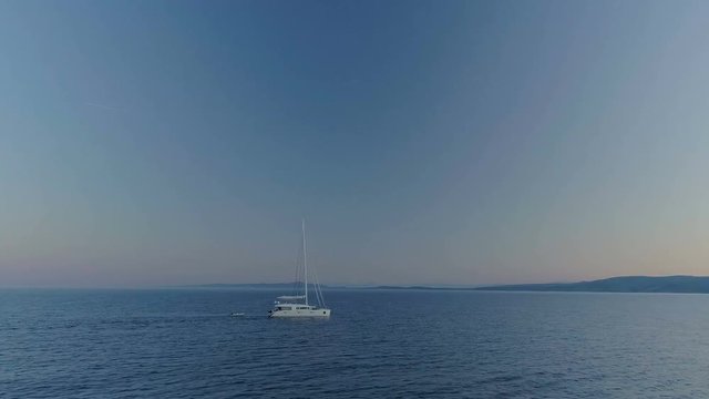 Aerial Time-Lapse Arc Shot of a White Sailing Yacht Traveling on a Calm Sea with Coast of Hills Visible. Early Morning/ Evening. Shot on Phantom 4K UHD Camera.