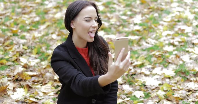 Attractive woman in black jacket taking selfie while sitting on grass covered with leaves during her walk in autumn park.