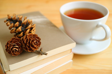 Cup of hot tea with dry pine cones on stack of books on natural color wooden table 