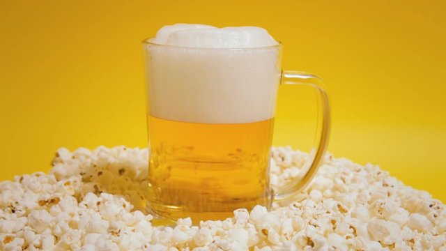 A glass filled with fresh golden beer and foam, rotating on a surface, together with a lot of popcorns. Yellow background.
