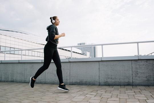 Young attractive lady jogging in the city near buildings. Beautiful happy girl running in the morning outside. Woman trains during the sunrise in the city center before breakfast. 