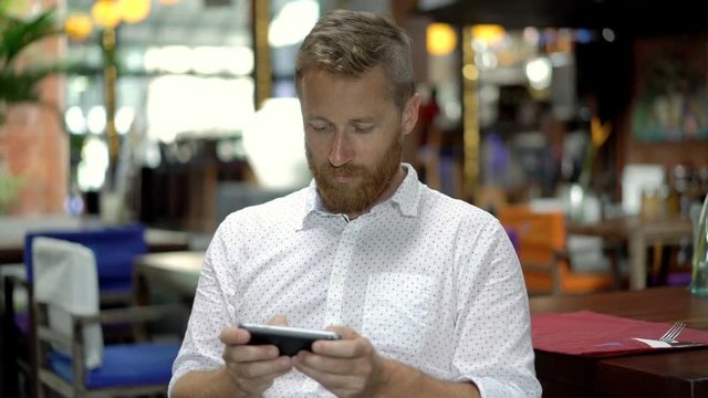 Absorbed man sitting in restaurant and browsing internet on smartphone, steadycam shot
