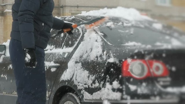 Caucasian man in casual clothes cleaning car from the snow near the house in winter against the backdrop of multi-storey residential building