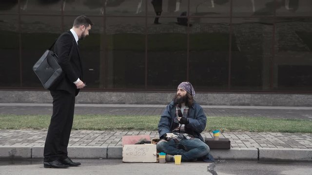The businessman banishes the bearded homeless man and throws back his cup with money.