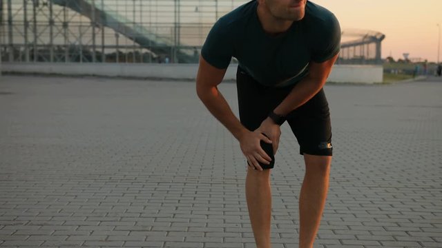 Close up. Portrait of a young runner feeling pain in his knee. Bending and holding his leg. Wearing a smartwatch. Blurred background.