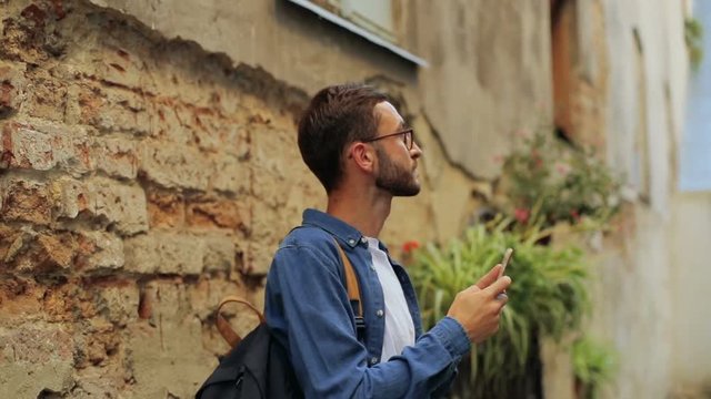 A portrait of a handsome young man walking along the old town and taking photos with his phone. Moving camera.