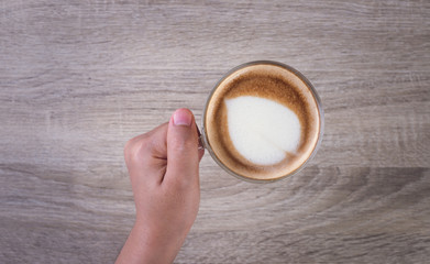 Female hands holding a cup of coffee with heart make up on face of milk cream on wooden table, top view