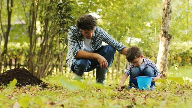 A portrait of a little boy and his dad gardening. Boy puts soil into his bucket. Dad taps him on his shoulder. Blurred background