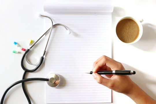 Hand Holding Pen in Flat lay photo with Notebook , Coffee Cup , Stethoscope ,Medical Capsuls on white background