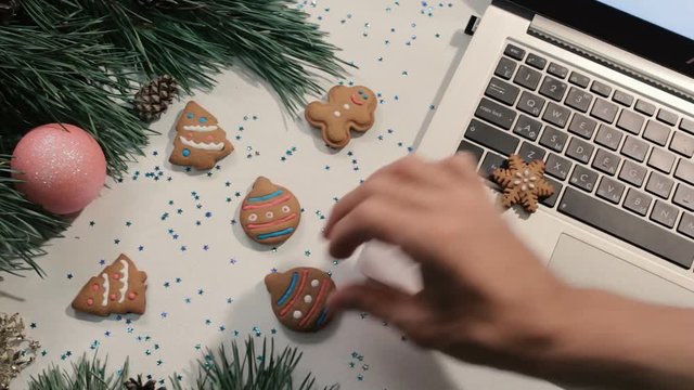 Cookies in Christmas and New Year holidays. Unrecognizable people lay sweet scones and gift on festive glittering table near decorated pine and laptop, top view. Concept of greetings at office