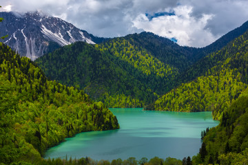 Fototapeta na wymiar Mountain lake surrounded by green forests on the slopes, incredible beauty against a background of melting snow on top. Ritsa lake, Relict national park of Ritsa, Abkhazia.