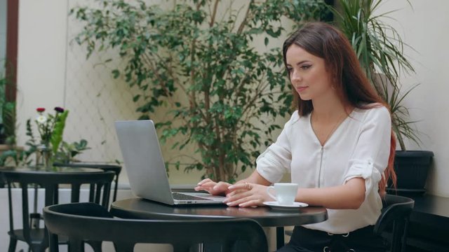 A beautiful girl sitting at the table in the pub and using her laptop. Medium shot. Soft focus.