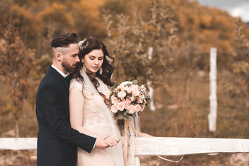 Stylish bride holding weadding bouquet standing with groom outdoors over autumn nature background....