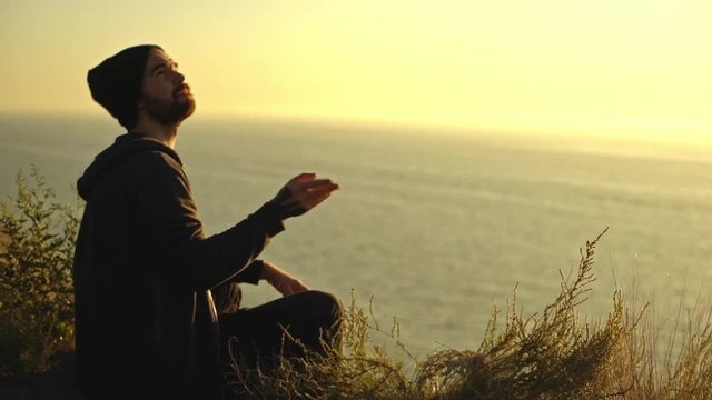 side view of caucasian guy wearing hat and hoodie sitting alone on the edge of hill over sea playing with footbag throwing it up at pleasant sunset slow motion
