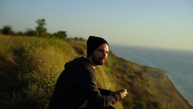 round footage of handsome bearded guy in hat sitting alone on hillside over sea playing with footbag throwing it up enjoying pastime in solitude slow motion